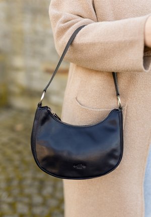 Borsa a spalla in pelle nera con cerniera indossata sul braccio sopra un cappotto beige, sfondo esterno sfocato.