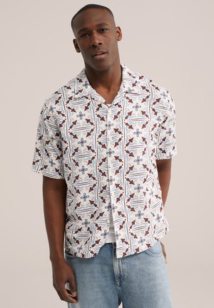 Homme portant une chemise à manches courtes avec un motif floral marron et bleu, et un jean bleu clair, debout devant un fond uni.