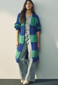 Lange gebreide cardigan in blauwe en groene strepen, gecombineerd met een grijze top en lichtblauwe wijde jeans. Toont geribbelde textuur en oversized pasvorm.