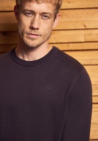 Dunkelvioletter Strickpullover mit Rundhalsausschnitt und dezentem Logo-Detail auf der linken Brust. Glatte Textur mit figurnahen Schnitt.