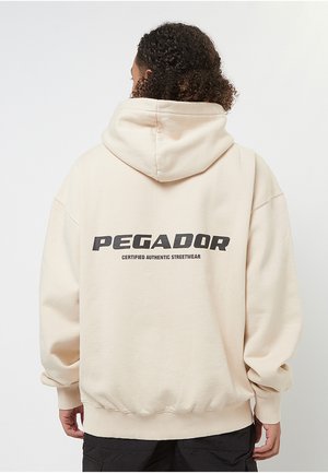 Persoană cu păr creț purtând un hanorac bej cu textul „PEGADOR CERTIFIED AUTHENTIC STREETWEAR” pe spate, stând în fața unui fundal simplu.