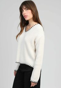 Pull en maille blanche avec un col en V, doté de bordures noires le long de l'encolure et de l'ourlet. Coupe ample avec des manches longues et une texture côtelée.