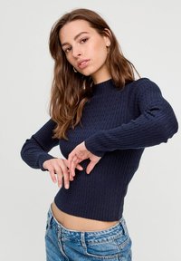 Granatowy sweter z prążkowanego materiału, z wysokim kołnierzem i długimi rękawami, w połączeniu z jasnoniebieskimi jeansami z efektem przetarcia. Widoczna faktura materiału.