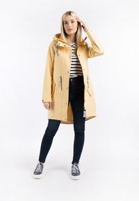 Un chubasquero amarillo impermeable con capucha, frente con cremallera, bolsillos y herrajes grises, combinado con una camisa a rayas y jeans oscuros.