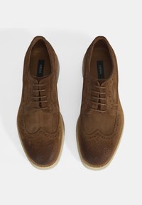 Chaussures en daim marron avec un design à bout ailé, perforations décoratives et une semelle en caoutchouc de couleur claire. Lacets et étiquette de marque visibles.