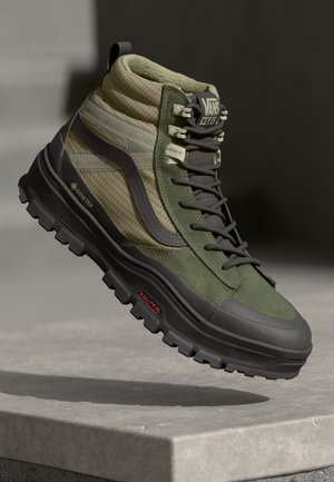 High-top green and beige Vans hiking boot with black chunky sole, Gore-Tex fabric, and all-terrain lugs, shown mid-air above a stone surface.
