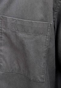 Pelēka kokvilnas krekls ar krūšu kabatu ar pogu, smalku diagonālu tekstūru un taisnu apakšmalu. Redzamas šuvju detaļas.