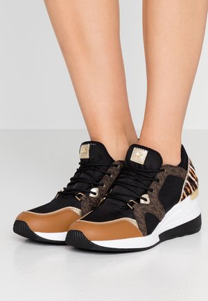 Voeten met zwarte, bruine en witte sneakers met dierenprintdetails en gouden accenten, staand op een lichtgrijze vloer.
