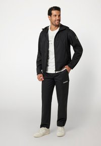 Juodas vandeniui atsparus striukė su gobtuvu, derinama su atitinkamais juodais kelniais, turinčiais prekės ženklą. Modelis vilki baltus sportinius batelius ir marškinėlius.