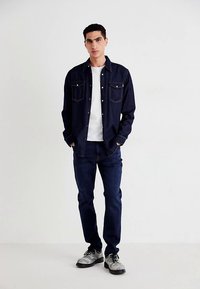 Tummansininen farkkupaita, jossa kaksi rintataskua, valkoisen t-paidan päällä, yhdistetty tummiin farkkuihin ja vaaleanharmaisiin kenkiin.