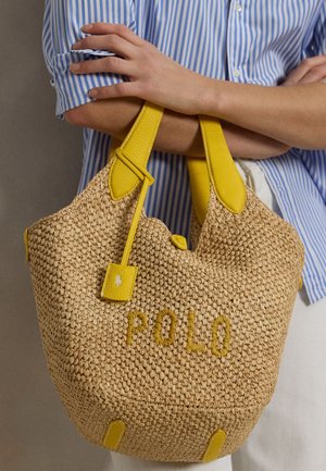 Persona con camisa azul a rayas sosteniendo un bolso tejido beige con asas de cuero amarillo y "POLO" bordado en el frente.
