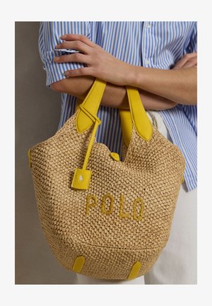 Persona con camicia a righe blu che tiene una borsa intrecciata beige con manici in pelle gialla e la scritta "POLO" ricamata sulla parte anteriore.
