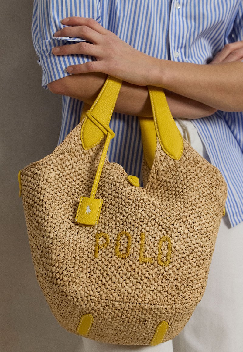 Personne en chemise rayée bleue tenant un sac à main beige tissé avec des poignées en cuir jaune et "POLO" brodé à l'avant.