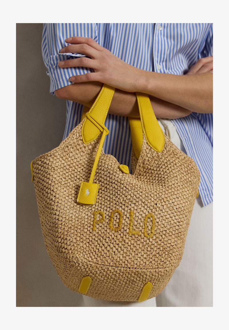 Osoba v modrej pruhovanej košeli drží tkanú béžovú kabelku so žltými koženými rukoväťami a výšivkou "POLO" na prednej strane.