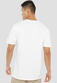 A man stands with his back facing the camera, wearing a plain white t-shirt and khaki pants, set against a gray background.