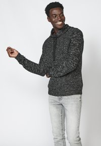 Maglione nero e bianco lavorato a maglia con cappuccio, caratterizzato da un motivo testurizzato e un laccio nero. Abbinato a jeans grigio chiaro.