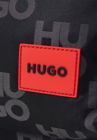 Fekete textúrájú táska, amelyen egy kiemelkedő, gumírozott piros címke található, rajta fekete betűkkel a "HUGO" felirattal, a szövet ismétlődő logójával háttérben.