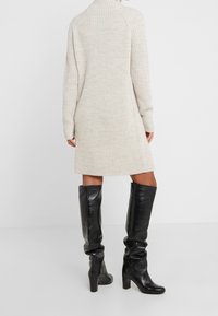 Persona vistiendo un vestido de punto acanalado color crema y botas de cuero negras de tacón alto hasta la rodilla, de pie contra un fondo blanco simple.