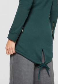 Sudadera larga de color teal con un dobladillo redondeado, cremalleras en los laterales y un cordón en la parte posterior. Tejido suave con una textura lisa.