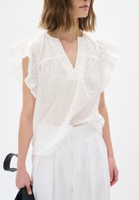 Vit blus med hålmönster, V-ringning och volangärmar. Matchad med vita veckade byxor och en svart handväska. Slät textur.