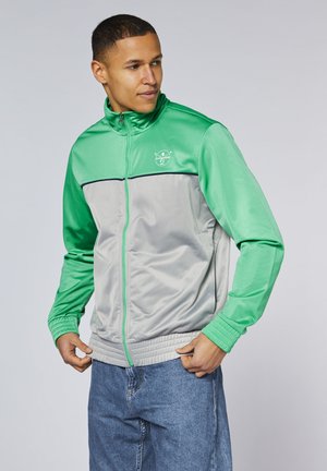 Ein Mann in einer grün-grauen Zip-Jacke und blauen Jeans steht nach links gewandt und hält den Jackensaum vor einem schlichten hellen Hintergrund.