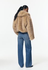 Femme à la queue de cheval noire portant une veste en fausse fourrure beige et un jean bleu à jambes larges avec des poches arrière cloutées, debout de profil.