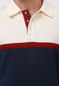 Polo majica s kremastim gornjim dijelom, crvenim ovratnikom i dugmadi, te tamnoplavim donjim dijelom, izrađena od teksturirane tkanine.
