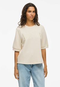 Helebež sweatshirt, millel on embosseeritud "AMOUR" tekst, lühikesed varrukad, ümar kaelus ja lõdva istuvusega. Koos siniste teksadega.