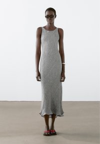 Grijze, mouwloze, tot de vloer reikende jurk met een gestructureerd ontwerp en gerimpelde zoom; gedragen met zwarte zonnebril en rode flipflops.