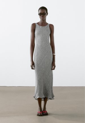 Grijze, gebreide, mouwloze jurk met een uitlopende zoom. Draag met zwarte zonnebril en rode sandalen. Soepele textuur en getailleerde silhouette.