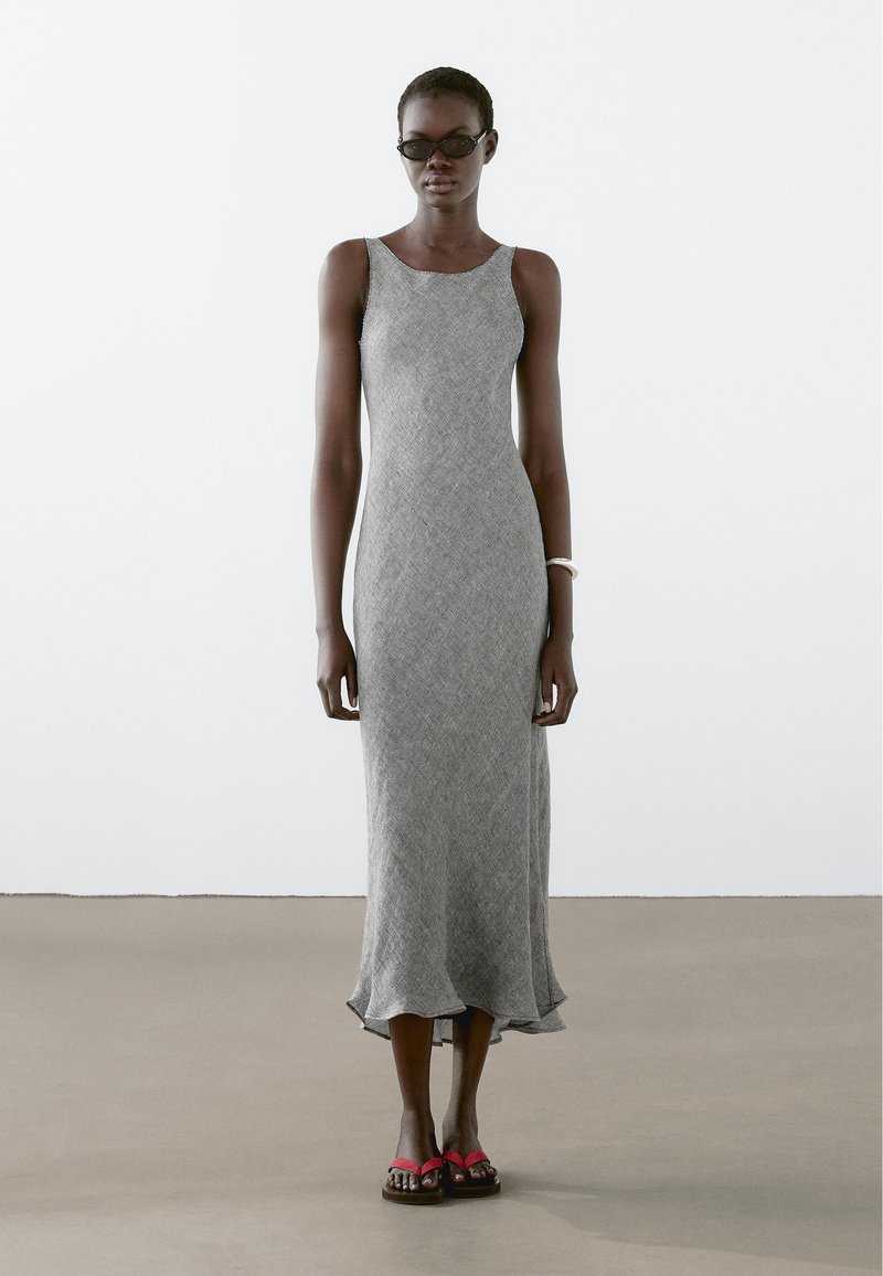 Grijze, mouwloze, tot de vloer reikende jurk met een gestructureerd ontwerp en gerimpelde zoom; gedragen met zwarte zonnebril en rode flipflops.