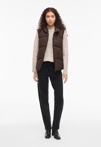 Pufasta smeđa prsluk s kapuljačom preko svijetlo bež džempera, uparen s crnim širokim hlačama i crnim gležnjačama. Jednostavna pozadina.