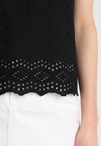 Black top featuring embroidered details and a scalloped hem with cut-out designs. Paired with white pants for contrast.