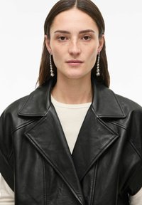 Veste noire en cuir à double boutonnage avec larges revers, finition texturée et coupe décontractée. Le modèle porte un haut crème et des boucles d'oreilles pendantes métalliques.