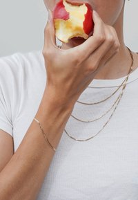 Gold layered necklaces with a delicate, flat chain design. A hand holding a partially eaten red apple, with smooth skin showing bright color.