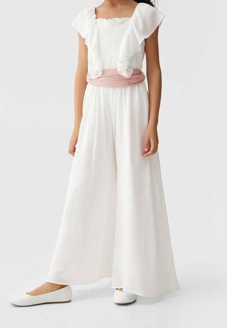 Jeune fille portant une combinaison sans manches blanche à volants avec une ceinture rose et des chaussures plates blanches.