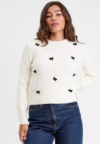 Pull en tricot blanc ornée d'appliqués en forme de nœud noir répartis uniformément sur le devant. Encolure classique en crew et poignets et ourlet côtelés.