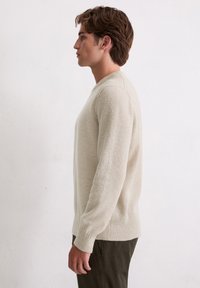 Jasno beżowy sweter dziany z ściągaczami na dole i rękawach, posiadający okrągły dekolt i miękkie, teksturowane wykończenie. Prezentowany na modelu męskim.