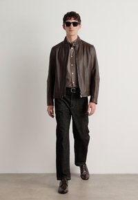 Veste en cuir marron à zip portée sur une chemise boutonnée marron clair, associée à un pantalon sombre et des chaussures marron. Des lunettes de soleil noires complètent le look.