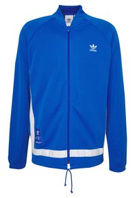 Sininen urheilutakki, jossa vetoketju edessä, korkea kaulus, valkoiset sivupaneelit ja kiristysnyöri helmassa. Valmistettu sileästä kankaasta, jossa Adidas-logo.