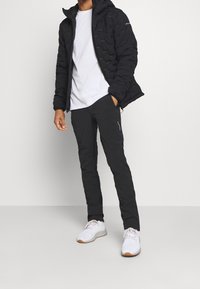 Svart vadderad jacka med en strukturerad yta, bärs över en vit t-shirt och svarta byxor, matchat med vita sneakers med ljusa detaljer.