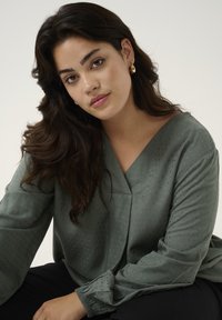 Green, textured blouse with a V-neck, long sleeves, and elastic cuffs. Fabric shows subtle patterns; color is soft and muted.