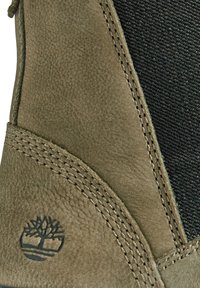 Gros plan sur une botte en cuir vert olive avec un panneau latéral en élastique noir et un logo Timberland en forme d'arbre embossé près de la semelle.