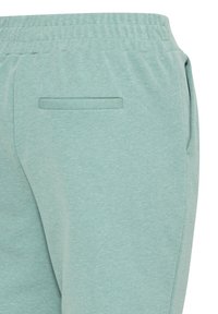 Primer plano de pantalones elásticos de cintura verde claro con un bolsillo trasero y detalles en las costuras laterales.