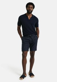 Marineblauw polo shirt met korte mouwen, gecombineerd met gestreepte marineblauwe shorts en tan sandalen met een zwarte band. Stijlvolle en casual zomerse outfit.