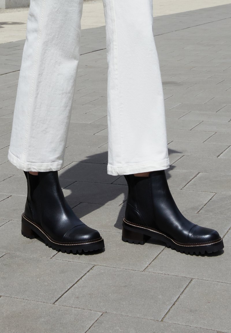 Personne portant un pantalon blanc et des bottes de cheville en cuir noir se tenant sur un trottoir gris clair à l'extérieur.