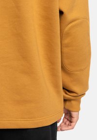 Sudadera mostaza con cuello redondo, puños de canalé y parches en los codos. Textura suave y diseño casual. Detalles de costura visibles en el dobladillo.