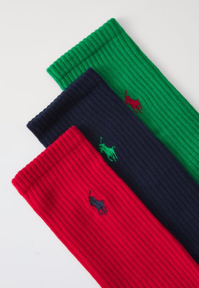 Trois paires de chaussettes côtelées en rouge, bleu marine et vert, chacune arborant un petit logo brodé. Les chaussettes sont en tissu épais et texturé.
