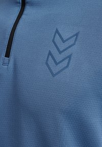 Blauwe textuurstof met zwarte ritssluiting en twee donkerblauwe chevron-logo's die verticaal aan de linkerkant zijn gerangschikt.