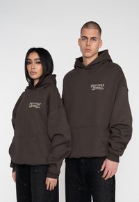 Felpe marroni con tasca frontale e logo, caratterizzate da una vestibilità comoda e polsini a coste, indossate da un uomo e una donna su uno sfondo neutro.